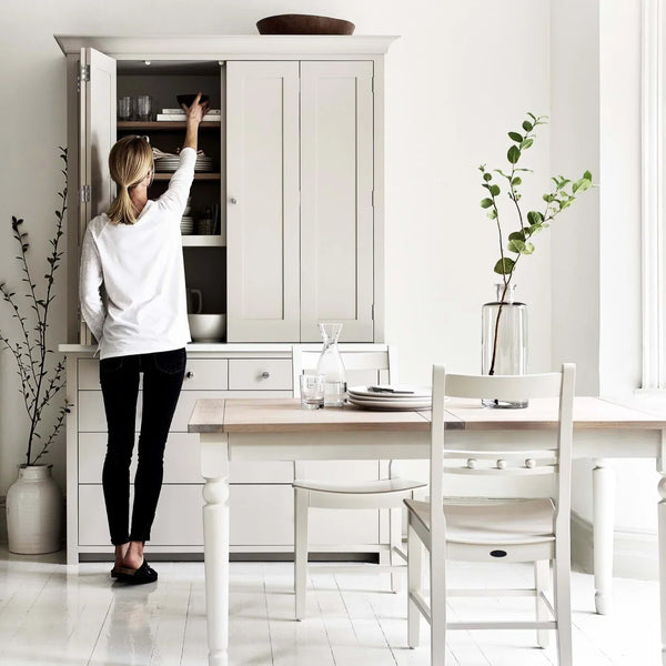 A woman in a white long-sleeve top and black pants reaches into the open upper cabinet of a large white kitchen hutch. The scene is a bright, modern dining room with a white table, chairs, and a vase with greenery.