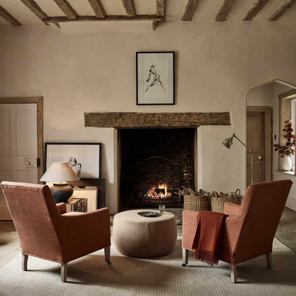 A cozy living room scene with two rust-orange upholstered armchairs facing a round beige ottoman, set in front of a fireplace with a wood beam above. The room has warm beige walls, rustic wooden ceiling beams, and framed wall art.