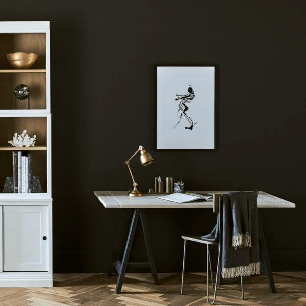 A minimalist home office scene featuring a black wall with a framed black and white line art print, a white desk with a marble top, a brass desk lamp, and a white shelving unit on the left.