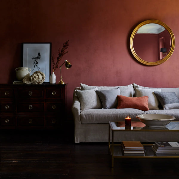 A styled living room corner featuring a dusty rose wall, a light gray sofa with gray and rust accent pillows, a gold-framed round mirror, a dark wood chest, and a glass coffee table.