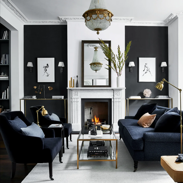 A styled living room interior with a white fireplace mantel, black accent walls, navy sofas and armchairs, gold-framed tables and lamps, framed wall art, a mirror, and a glass coffee table in the center.
