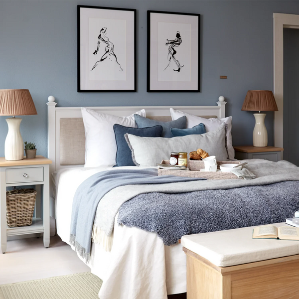 A styled bedroom scene with a neatly made bed featuring white and blue bedding, multiple pillows, a blue throw blanket, and a wooden bench at the foot. Two matching nightstands with lamps flank the bed, and two framed abstract black line drawings hang on the blue wall above.