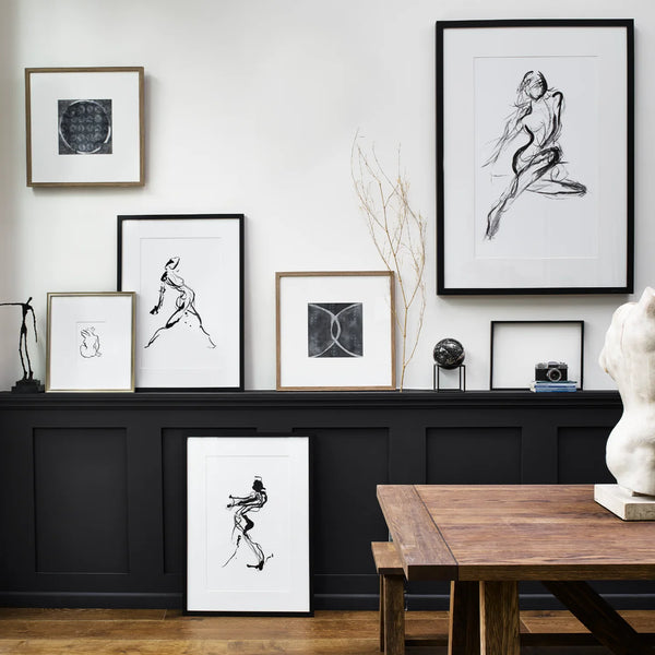 A styled interior wall display featuring multiple framed black-and-white art prints in different frame finishes, arranged above a dark wood console and next to a wooden table with a white sculpture.