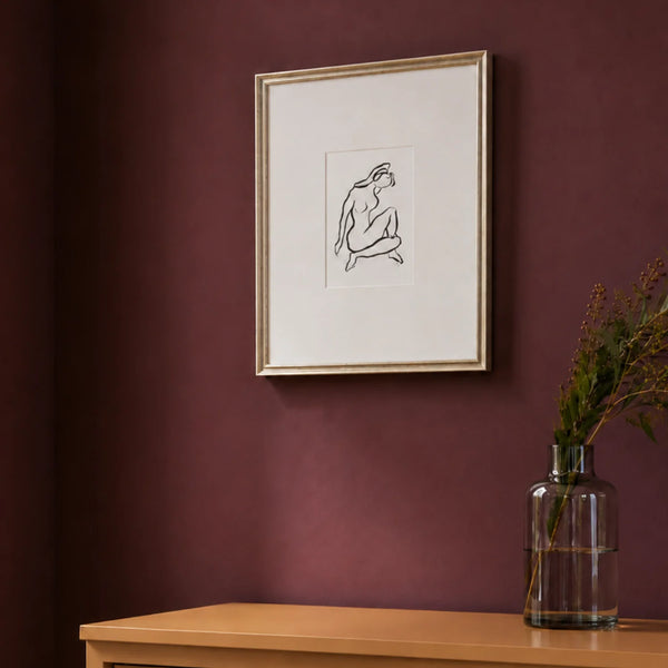 A framed minimalist black line art print of a seated figure on a white mat, hanging on a dark burgundy wall above a light wood console table with a glass vase containing dried stems.