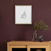 A framed minimalist line art print of a seated figure, displayed on a dark plum wall above a light wood console table with a glass vase and decorative object.