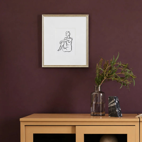 A framed minimalist line art print of a seated figure, displayed on a dark plum wall above a light wood console table with a glass vase and decorative object.