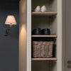 An open interior closet or shelf unit with light wood shelves, containing a pair of white sneakers on the top shelf, black shoes on the middle shelf, and a woven wicker basket on the bottom shelf. A wall-mounted lamp with a beige shade is visible on the left against a dark gray paneled wall.