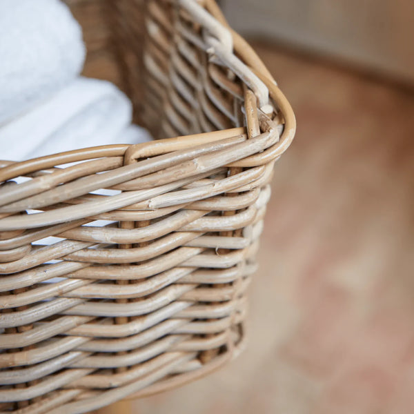 A close-up of a natural woven wicker basket containing folded white towels, set against a softly blurred warm-toned indoor background.