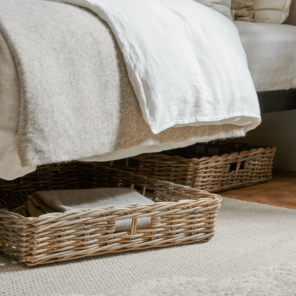 Natural woven wicker storage baskets placed underneath a bed with neutral-toned bedding and a white throw blanket, set on a light rug in a bedroom setting.