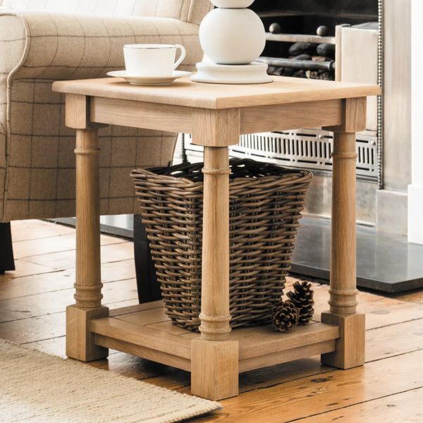 A light natural wood end table with turned legs and a lower shelf, styled in a living room setting with a wicker basket, a white ceramic vase, a teacup, and a pinecone on a wooden floor.