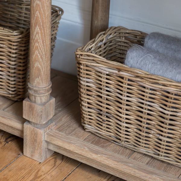A close-up of a natural wicker basket placed on a wooden shelf next to a wooden table leg, with folded gray towels inside the basket.