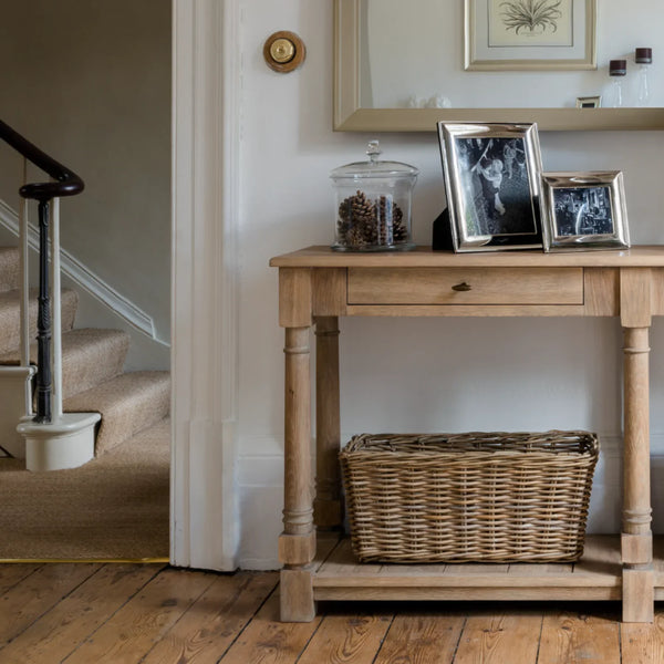 A light natural wood console table with a single drawer and lower shelf, styled with picture frames, a glass jar, and a woven wicker basket in a home entryway.
