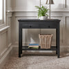 A black wooden console table with two drawers and an open lower shelf, styled in a home entryway with a potted fern, lamp, basket, and books. The table is set on a textured beige rug against a beige paneled wall.