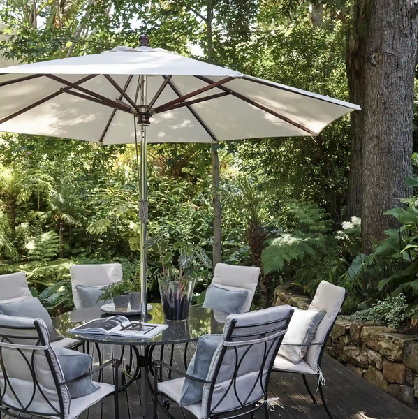 A large cream patio umbrella with a dark metal frame shading a glass-top outdoor dining table and four cushioned chairs on a wooden deck, surrounded by lush green garden foliage.