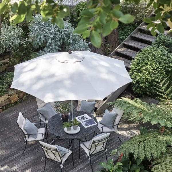 A large white patio umbrella shades a round outdoor dining table with six chairs in a lush garden courtyard.