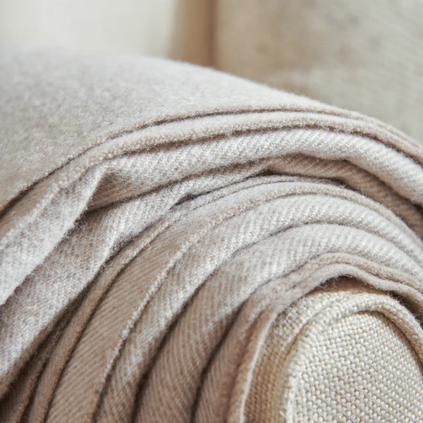 Close up of a folded cream-colored throw blanket with fringe trim draped over the arm of a matching upholstered chair