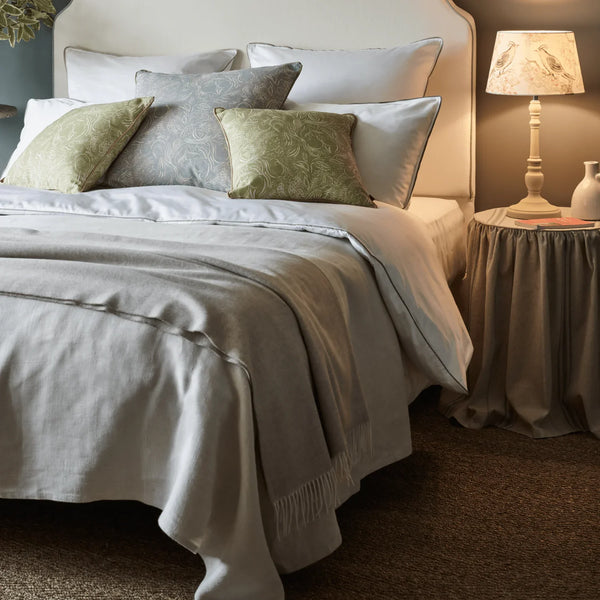 A styled bed in a bedroom setting with light gray and white bedding, two olive green patterned accent pillows, a beige upholstered headboard, a bedside table with a lit lamp, and brown carpet.