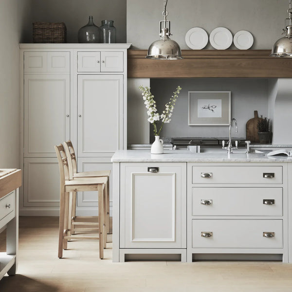 A styled kitchen interior featuring white shaker-style cabinetry and island with light wood countertops and stools, silver pendant lights, marble countertops, and a gray backsplash.