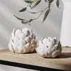 Two white ceramic artichoke-shaped candle holders on a light wood surface with a neutral background and soft shadows.