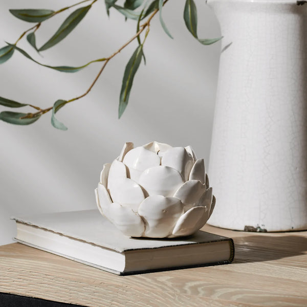 White ceramic artichoke-shaped candle holder on a light wood surface with a neutral background and soft shadows.
