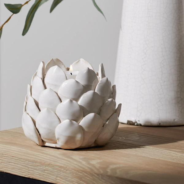 A white ceramic candle holder shaped like a lotus flower, lit with a small candle, resting on a light wood surface next to a textured white vase.