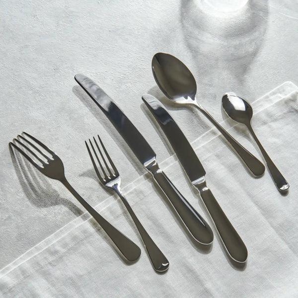 A six-piece stainless steel flatware set arranged diagonally on a textured white linen napkin. The set includes two dinner forks, two dinner knives, a dinner spoon, and a soup spoon, all reflecting soft light.