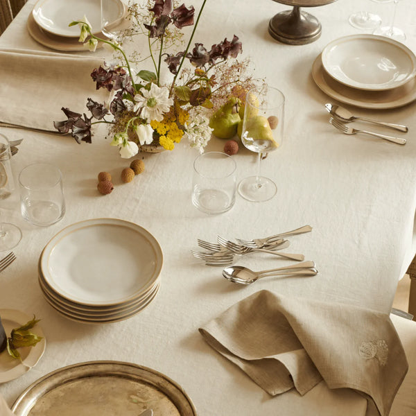 A styled dining table setting with cream linen tablecloth, stacked white plates with gold trim, silver cutlery, clear drinking glasses, folded beige napkins, and a floral centerpiece with green apples and small dried botanicals.