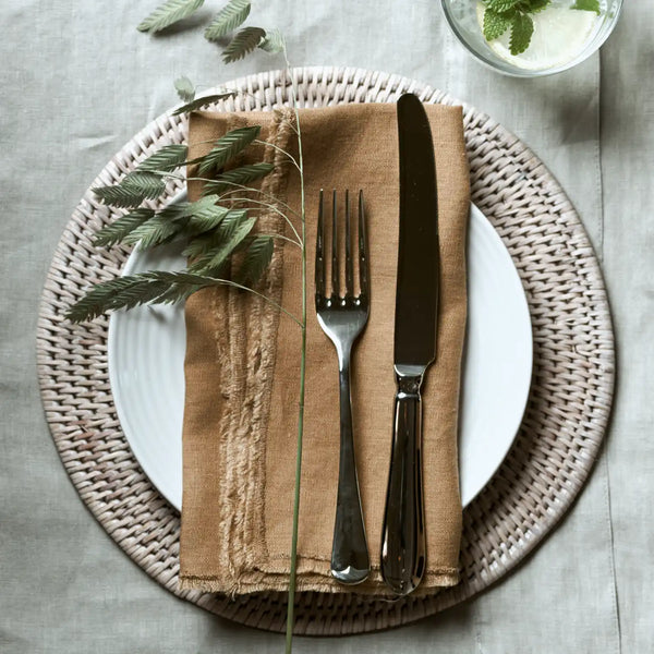 A styled table setting featuring a white ceramic plate on a round woven placemat, with a folded tan linen napkin holding a silver fork and knife, and dried green stems on a light gray linen tablecloth.