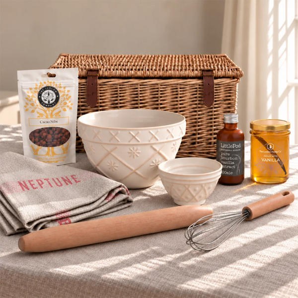 A styled baking or cooking kit arranged on a tablecloth with warm natural light, including a wicker basket, cream ceramic bowls, a wooden rolling pin, metal whisk, vanilla extract bottle, cocoa nibs pouch, and a jar of vanilla extract.