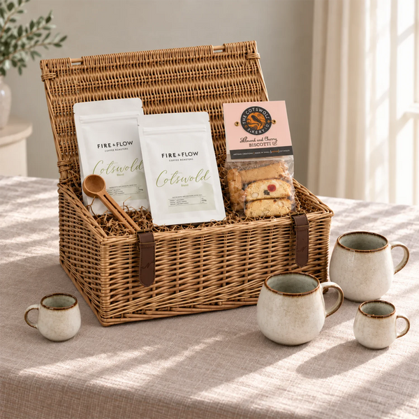 A wicker picnic basket filled with FIRE+FLOW Cotswold coffee, a bag of biscuits, and a wooden spoon, set on a table with matching ceramic mugs in a softly lit indoor setting.