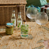 A styled picnic-style product arrangement on a woven blanket with a wicker basket, two clear textured wine glasses, a jar of olive and pistou, and three bottles including a clear bottle labeled 'OLIO sister' and a green-labeled bottle, with wrapped gifts in the background.