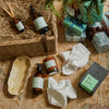 A styled flat-lay of wellness and home products arranged on a woven textile with pine branches and wrapped gifts. Visible items include amber bottles, a candle, white towels, a ceramic dish, and a wicker basket with a reed diffuser bottle.