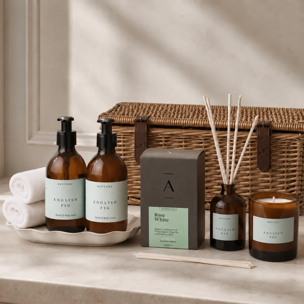A curated spa gift set arranged on a stone surface in front of a wicker basket. The set includes two amber glass hand wash bottles with black pumps and pale green labels, a gray box labeled 'Rose White', a reed diffuser bottle with wooden reeds, a candle jar, two rolled white towels, and two wooden sticks.
