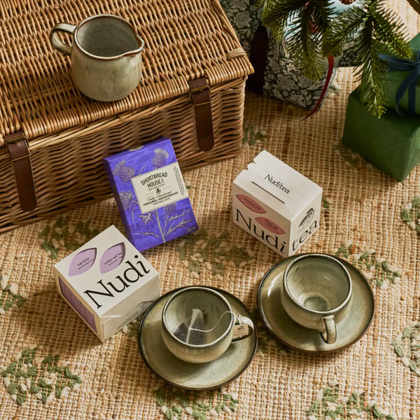 A styled flat-lay of Nudi tea products on a woven beige tablecloth, featuring two white tea boxes with purple and pink labels, a purple herbal tea box, and two ceramic teacups and saucers next to a wicker picnic basket.