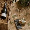 A curated gift basket arrangement on a burlap surface with a wicker basket, a bottle of Bolney's Port wine, two clear textured wine glasses, a tin of almonds, and Montezuma Absolute Black chocolate.