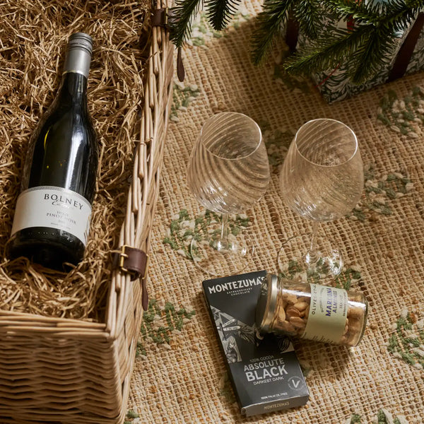 A curated gift basket arrangement on a burlap surface with a wicker basket, a bottle of Bolney's Port wine, two clear textured wine glasses, a tin of almonds, and Montezuma Absolute Black chocolate.