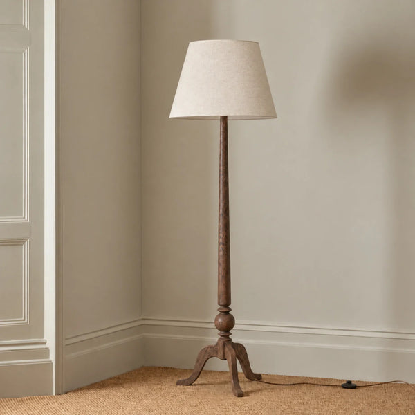 A rustic wooden floor lamp with a tapered beige fabric shade stands on a woven rug in a corner room with beige walls and white trim.