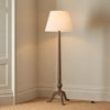 A classic floor lamp with a dark natural wood tripod base and a tapered beige fabric drum shade, shown lit in a corner of a room with beige walls and carpet.