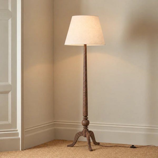 A classic floor lamp with a dark natural wood tripod base and a tapered beige fabric drum shade, shown lit in a corner of a room with beige walls and carpet.