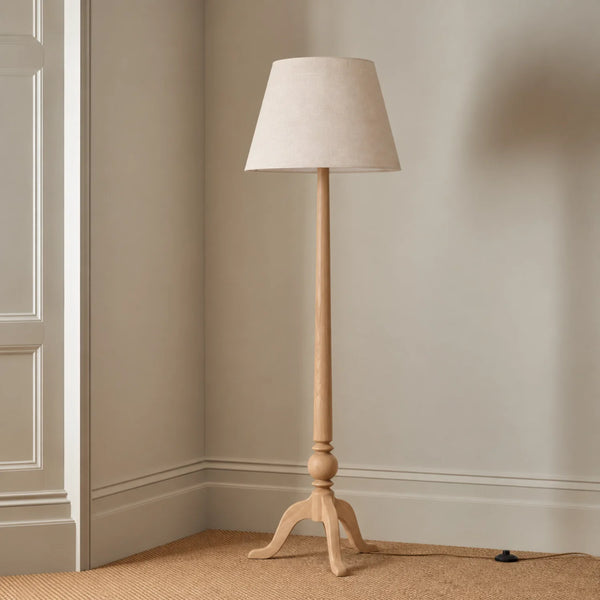 A classic floor lamp with a light natural wood tripod base and a tapered beige fabric drum shade, shown in a corner of a room with beige walls and carpet.