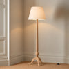 A classic floor lamp with a light natural wood tripod base and a tapered beige fabric drum shade, shown lit in a corner of a room with beige walls and carpet.