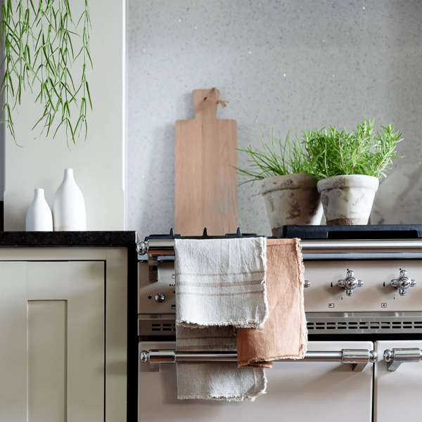 Three textured linen kitchen towels in cream, beige, and light brown tones are draped over the handles of a cream-colored vintage-style range stove in a kitchen setting.