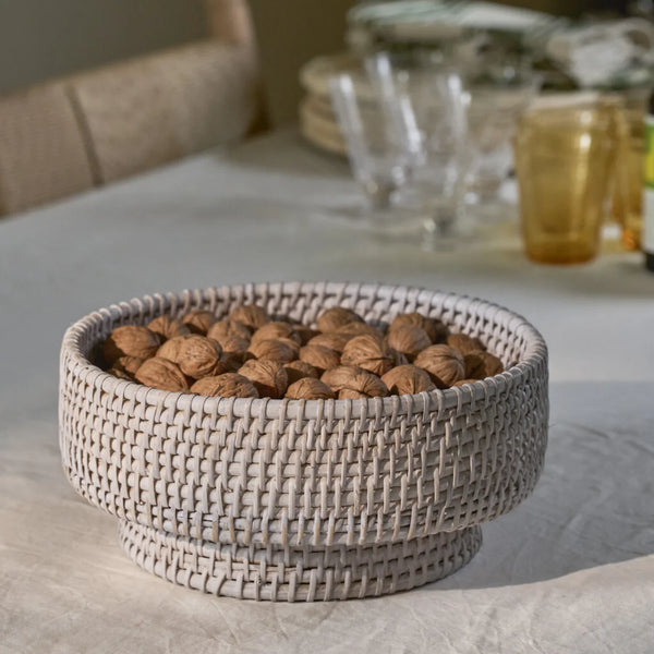 A round woven basket bowl filled with walnuts, set on a light beige tablecloth with a softly blurred dining table background.