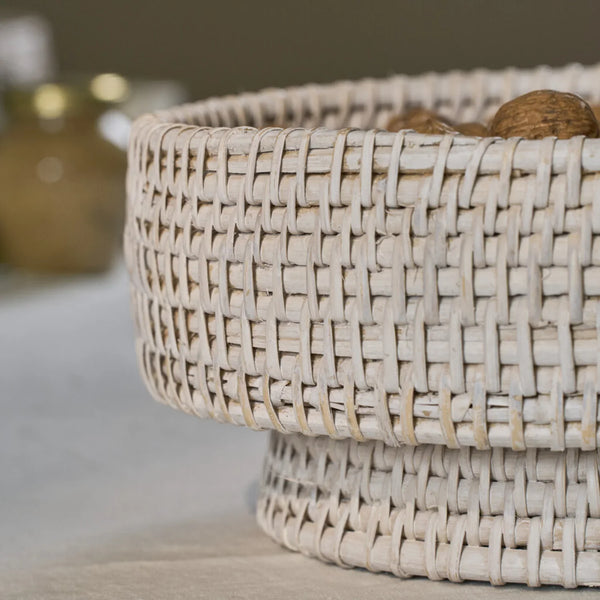 A close-up of a round woven basket made from natural light-colored wicker, shown sitting on a neutral surface with a softly blurred background.