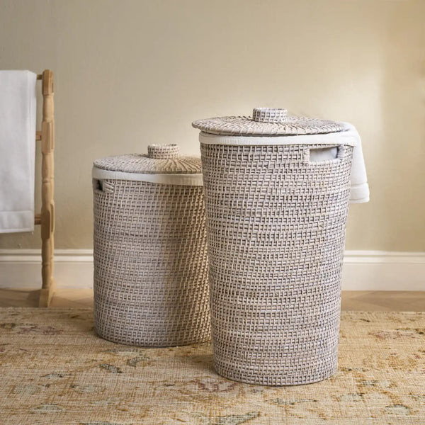 Two woven cylindrical laundry hampers in different sizes with matching lids, styled on a patterned rug against a beige wall.