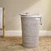 A woven cylindrical laundry hamper with a matching lid, shown in a real interior setting on a patterned rug against a beige wall.