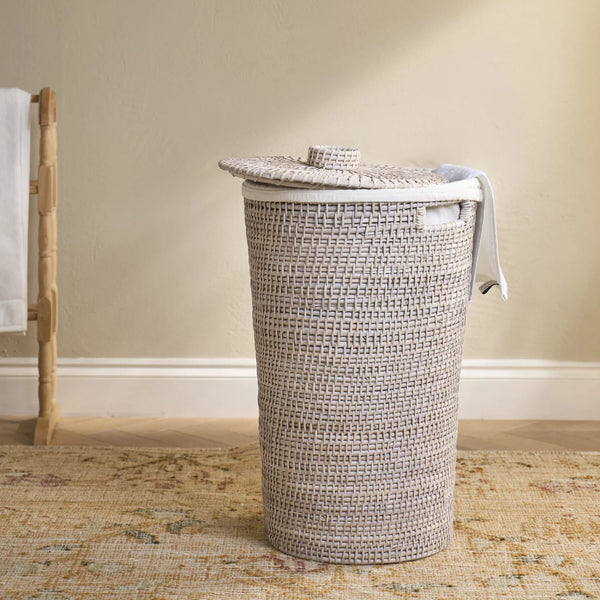A woven cylindrical laundry hamper with a matching lid, shown in a real interior setting on a patterned rug against a beige wall.