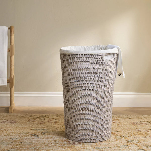 A tall woven cylindrical laundry hamper in a light gray and white basket weave, lined with a white fabric liner and shown standing on a patterned rug against a beige wall.