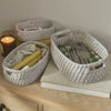 Three oval woven storage baskets in a light natural fiber material, styled on a light wood dresser with a candle, book, and tableware inside.