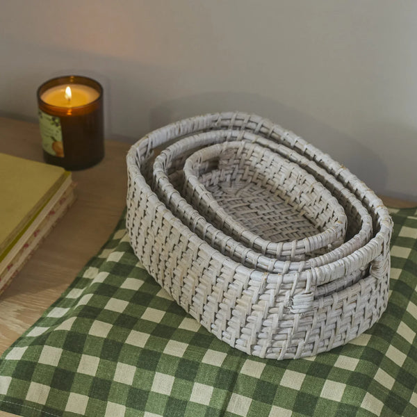 A set of nested oval woven baskets in a light gray color, styled on a green and white checkered blanket next to a lit candle and books.